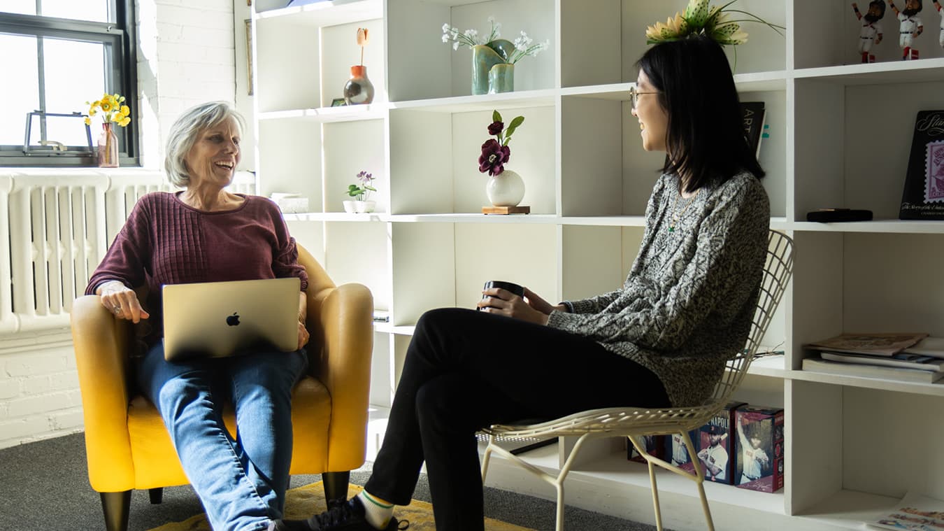 Two coworkers at RLMG sitting in chairs in an office discussing work while smiling